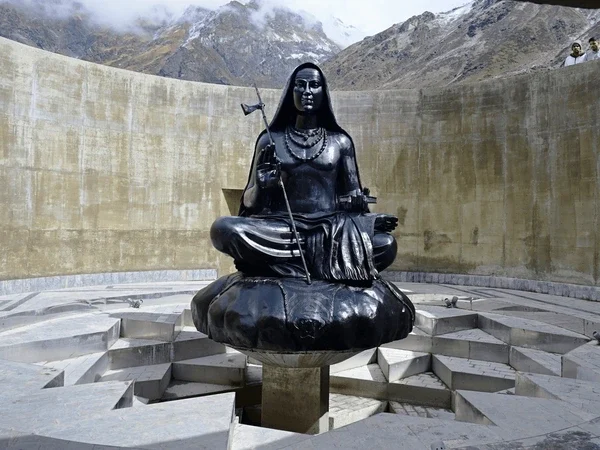 A black stone statue of Adi Shankaracharya seated cross-legged on a carved lotus pedestal inside a circular walled enclosure at Kedarnath, with snow-dusted peaks visible over the wall